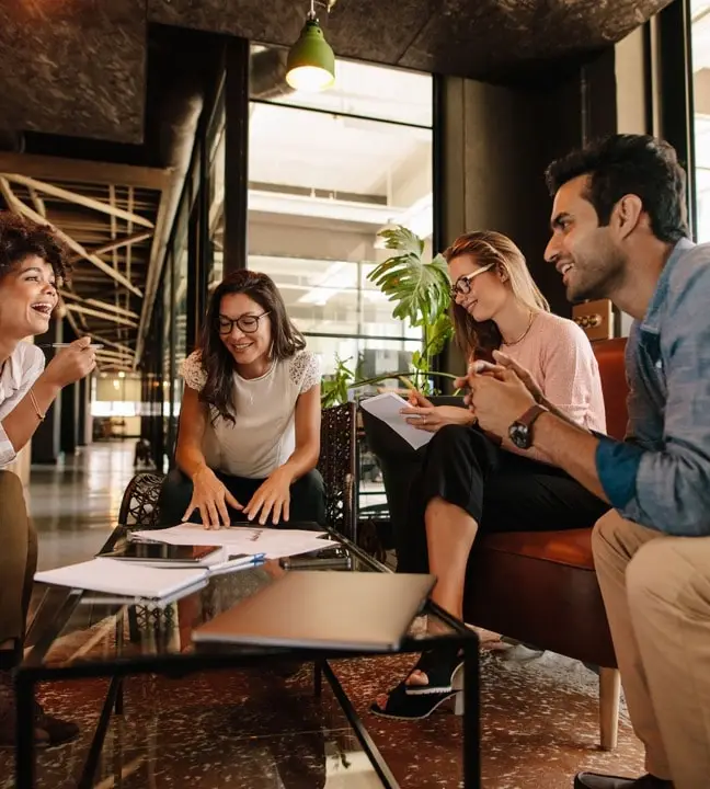 A group of four people sits around a coffee table in a modern office, engaged in a lively discussion. Papers and a laptop are on the table. The setting is light and airy, with a large plant in the background.